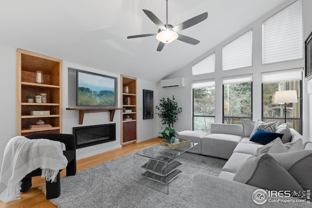 living area with built in features, a wall unit AC, wood finished floors, a glass covered fireplace, and high vaulted ceiling