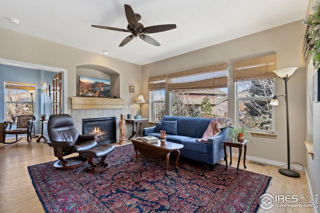 living area featuring visible vents, a ceiling fan, a tiled fireplace, wood finished floors, and baseboards