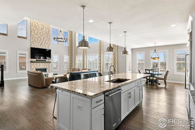 kitchen with an island with sink, dark wood-style flooring, a tile fireplace, stainless steel dishwasher, and a sink