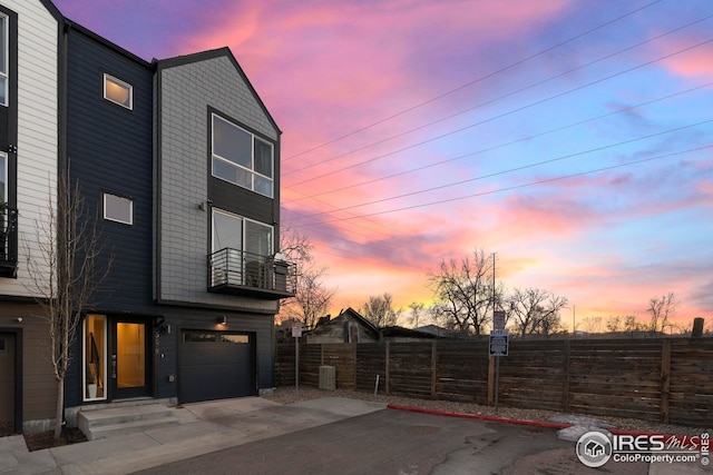 property exterior at dusk with a balcony, an attached garage, driveway, and fence