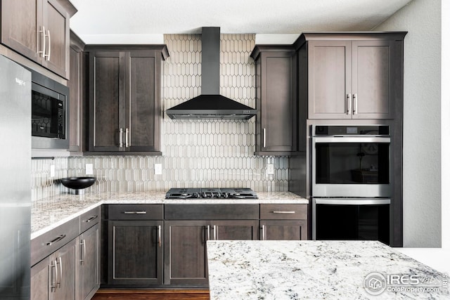 kitchen featuring light stone counters, tasteful backsplash, appliances with stainless steel finishes, wall chimney range hood, and dark brown cabinets