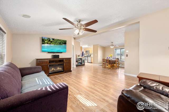 living area featuring a glass covered fireplace, a textured ceiling, light wood-style floors, and ceiling fan