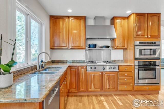 kitchen with light stone countertops, a sink, appliances with stainless steel finishes, wall chimney exhaust hood, and light wood-type flooring