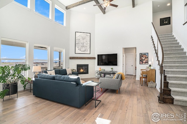 living area with beam ceiling, ceiling fan, stairs, a glass covered fireplace, and light wood-type flooring