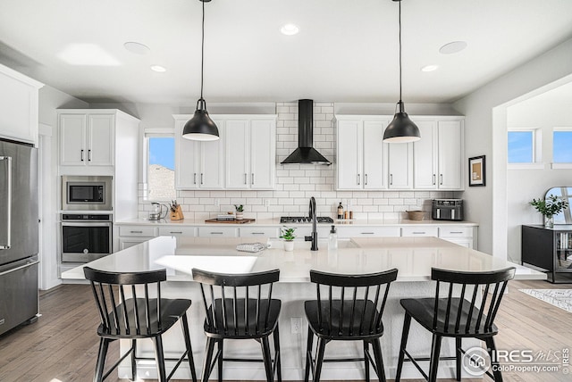 kitchen featuring backsplash, wall chimney range hood, light countertops, light wood-style floors, and stainless steel appliances
