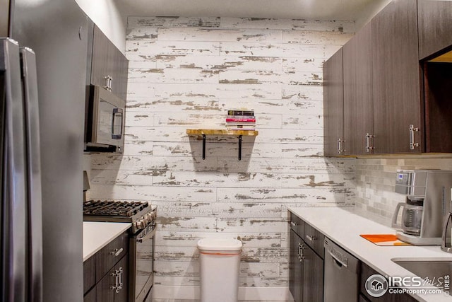 kitchen featuring wallpapered walls, stainless steel appliances, light countertops, dark brown cabinetry, and backsplash