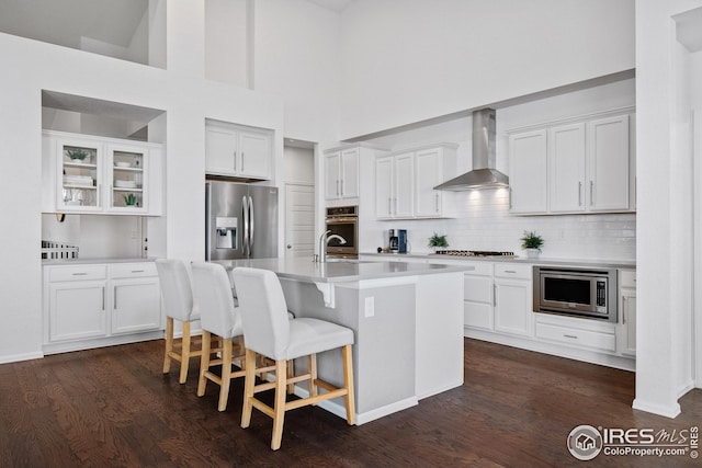 kitchen with a breakfast bar area, stainless steel appliances, an island with sink, wall chimney range hood, and white cabinets