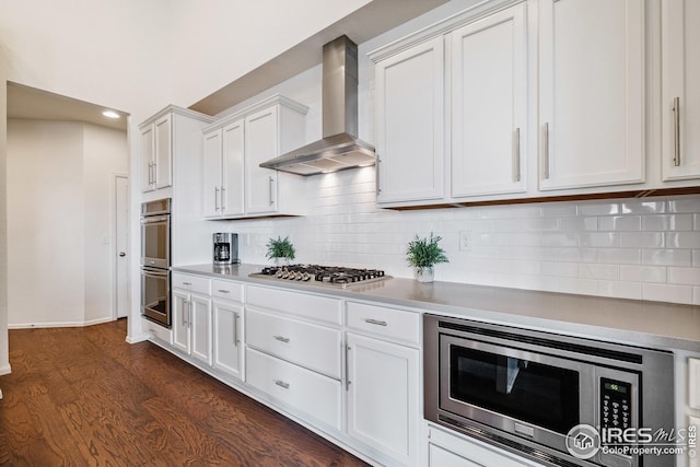 kitchen featuring decorative backsplash, dark wood-style flooring, white cabinets, appliances with stainless steel finishes, and wall chimney range hood