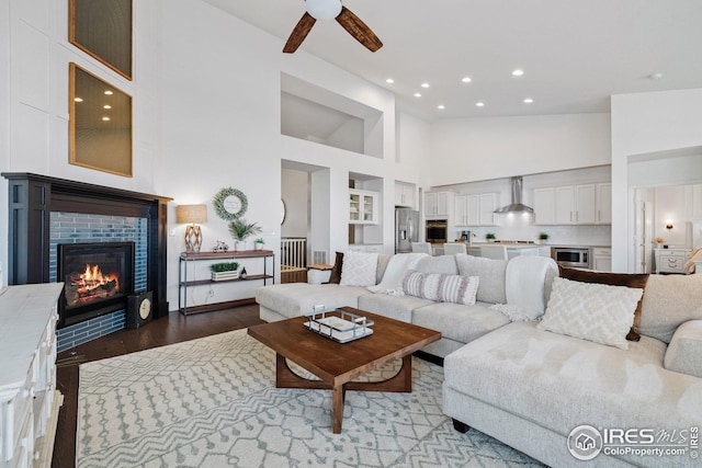 living area featuring a ceiling fan, wood finished floors, a glass covered fireplace, a towering ceiling, and recessed lighting