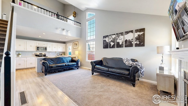 living room featuring high vaulted ceiling, stairway, light wood-style flooring, baseboards, and visible vents