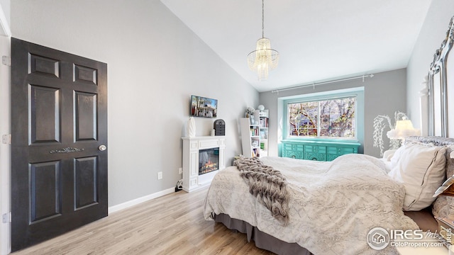 bedroom with vaulted ceiling, a glass covered fireplace, light wood-type flooring, baseboards, and an inviting chandelier