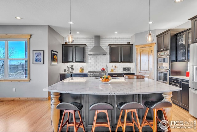 kitchen with wall chimney range hood, light stone counters, light wood-style floors, stainless steel appliances, and backsplash