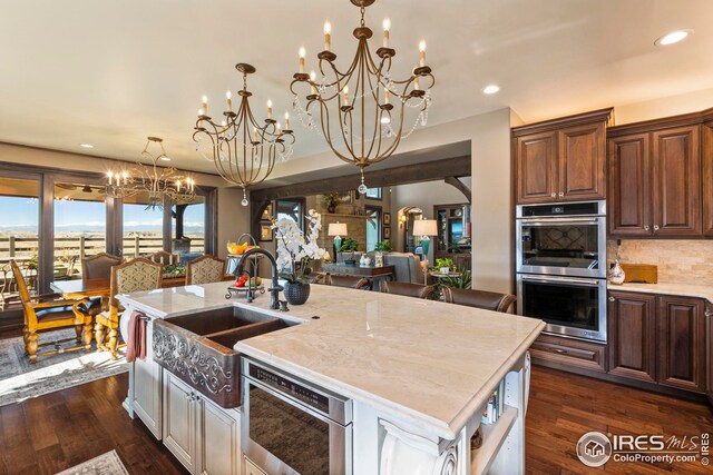 kitchen with a notable chandelier, a sink, stainless steel double oven, a center island with sink, and dark wood-style floors