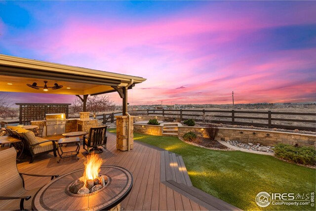 deck at dusk with a fenced backyard, an outdoor fire pit, area for grilling, and a yard