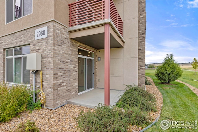 property entrance with stucco siding, brick siding, a balcony, a patio, and a yard