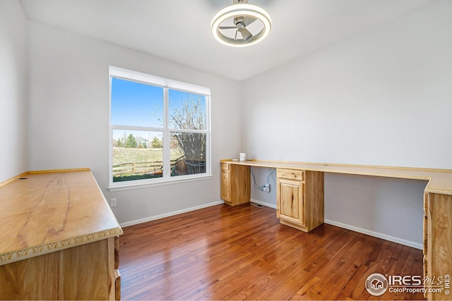 unfurnished office featuring built in desk, dark wood finished floors, baseboards, and ceiling fan