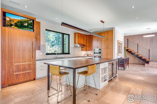 kitchen featuring tasteful backsplash, under cabinet range hood, a sink, dark countertops, and stainless steel double oven