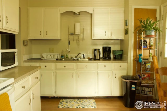 kitchen with white microwave, light countertops, white cabinetry, a sink, and wood finished floors