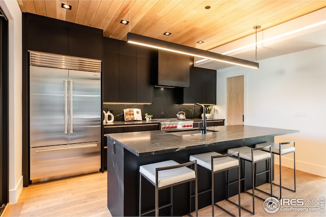 kitchen with built in refrigerator, a sink, a breakfast bar, dark cabinetry, and wooden ceiling
