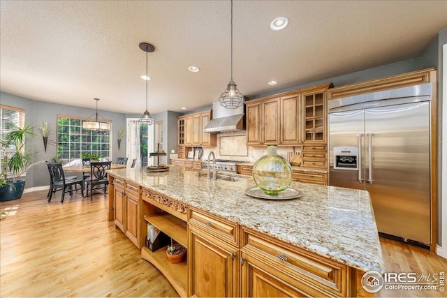 kitchen with open shelves, wall chimney exhaust hood, brown cabinets, stainless steel built in fridge, and a sink