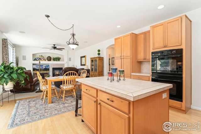 kitchen with arched walkways, dobule oven black, light wood-style floors, a ceiling fan, and tile counters