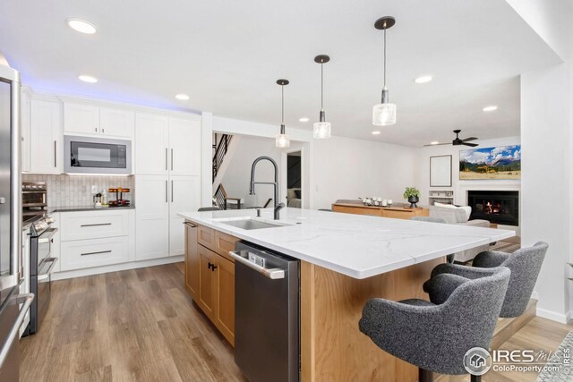 kitchen with stainless steel appliances, a sink, a center island with sink, light wood-style flooring, and ceiling fan
