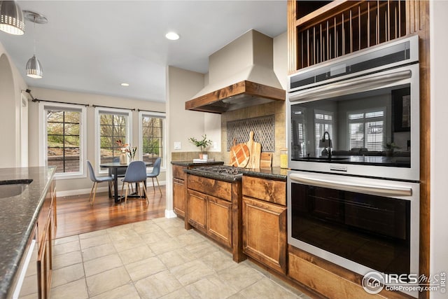 kitchen featuring dark stone countertops, hanging light fixtures, brown cabinets, exhaust hood, and appliances with stainless steel finishes