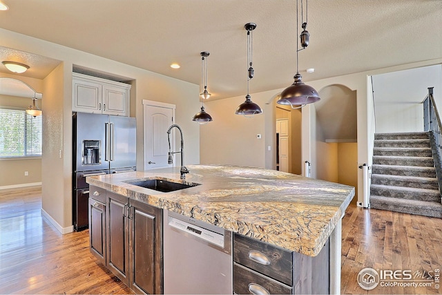 kitchen with light wood finished floors, a kitchen island with sink, a sink, baseboards, and appliances with stainless steel finishes