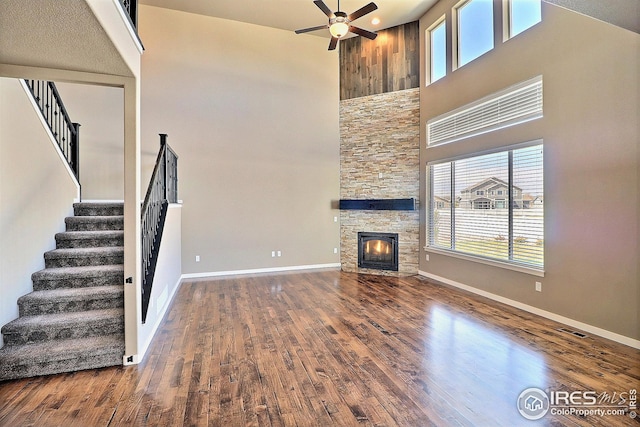 unfurnished living room with ceiling fan, baseboards, a towering ceiling, a stone fireplace, and stairs