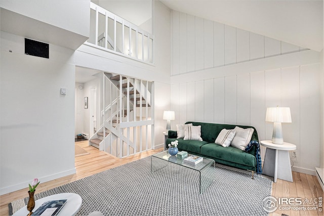 living room featuring a towering ceiling, light wood-style floors, baseboards, baseboard heating, and stairway