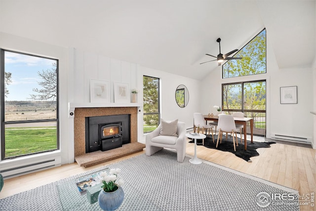 living area with a warm lit fireplace, ceiling fan, high vaulted ceiling, a baseboard radiator, and hardwood / wood-style flooring