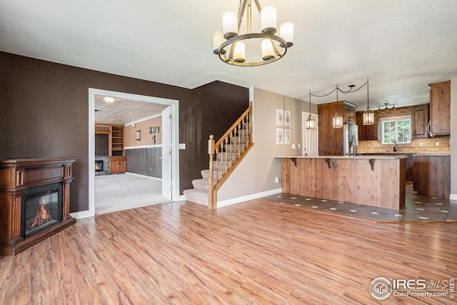 unfurnished living room featuring a glass covered fireplace, baseboards, stairway, light wood finished floors, and a notable chandelier