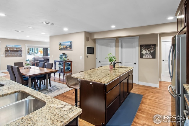 kitchen with freestanding refrigerator, dark brown cabinetry, an island with sink, a sink, and light wood-style flooring