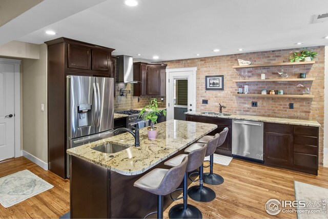 kitchen with dark brown cabinets, wall chimney range hood, a sink, light wood finished floors, and stainless steel appliances