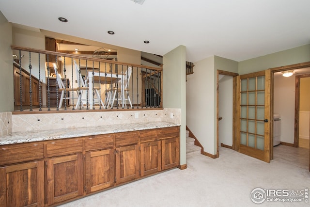 kitchen featuring brown cabinetry, light carpet, light stone countertops, baseboards, and tasteful backsplash