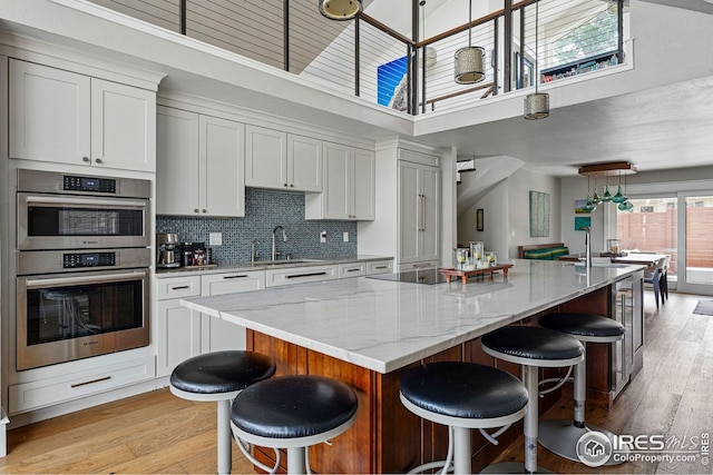 kitchen featuring stainless steel double oven, a sink, light wood finished floors, and a towering ceiling