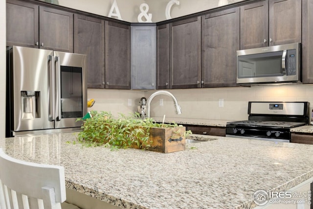 kitchen featuring dark brown cabinetry, a kitchen island with sink, tasteful backsplash, light stone counters, and stainless steel appliances