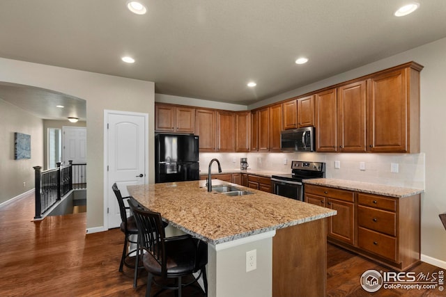 kitchen with brown cabinetry, appliances with stainless steel finishes, a sink, light stone counters, and dark wood-style floors