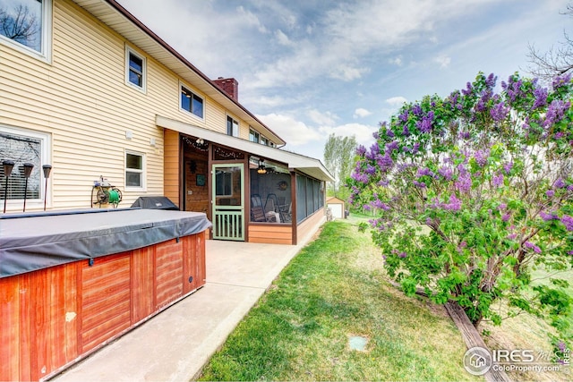 back of property featuring a hot tub, a sunroom, a chimney, a patio area, and a lawn