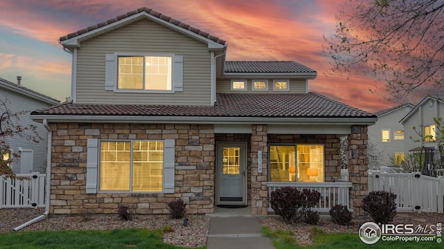traditional-style home with a tiled roof, fence, covered porch, and stone siding