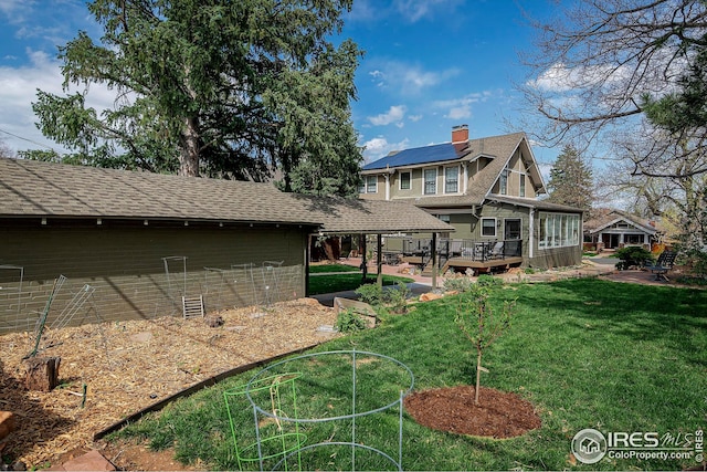 rear view of house featuring a chimney, a lawn, a wooden deck, roof mounted solar panels, and a shingled roof