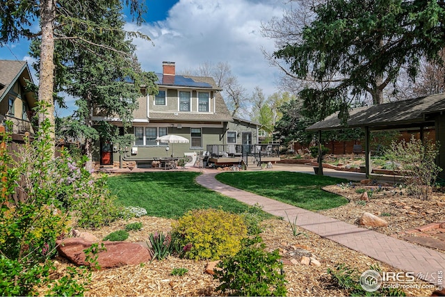 back of house featuring a wooden deck, a chimney, solar panels, a patio area, and a yard