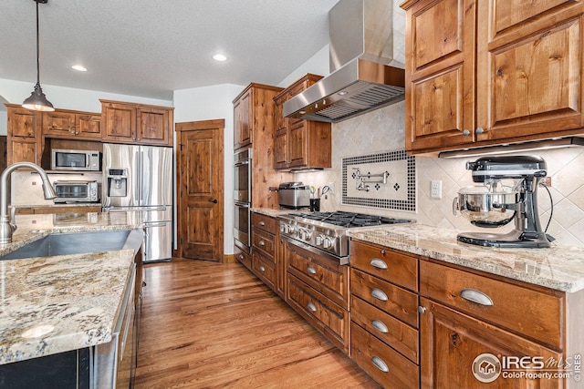 kitchen with wall chimney range hood, brown cabinets, a sink, stainless steel appliances, and light wood-type flooring
