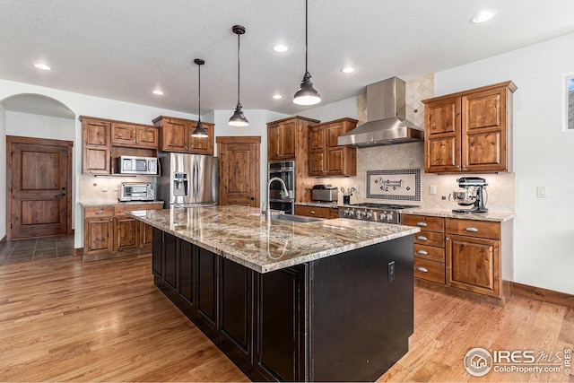kitchen featuring wall chimney range hood, brown cabinets, appliances with stainless steel finishes, light wood finished floors, and arched walkways