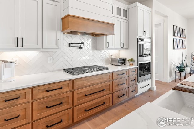 kitchen with premium range hood, white cabinets, tasteful backsplash, light wood-style flooring, and appliances with stainless steel finishes