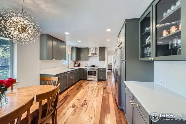 kitchen with wall chimney range hood, appliances with stainless steel finishes, light wood-type flooring, a sink, and light countertops