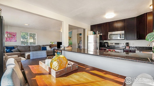 kitchen with appliances with stainless steel finishes, dark brown cabinets, dark stone countertops, a sink, and a peninsula