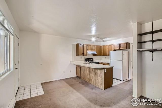 kitchen featuring freestanding refrigerator, under cabinet range hood, range, a peninsula, and brown cabinets