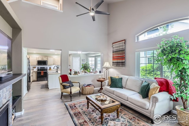 living area with a ceiling fan, light wood-type flooring, a high ceiling, plenty of natural light, and a stone fireplace