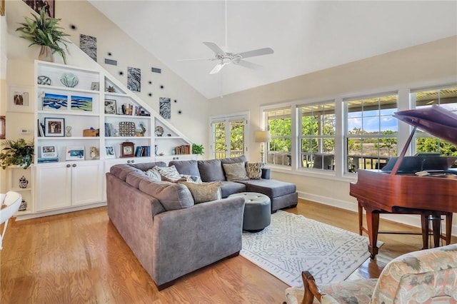 living room featuring built in shelves, light wood-style floors, a ceiling fan, high vaulted ceiling, and baseboards